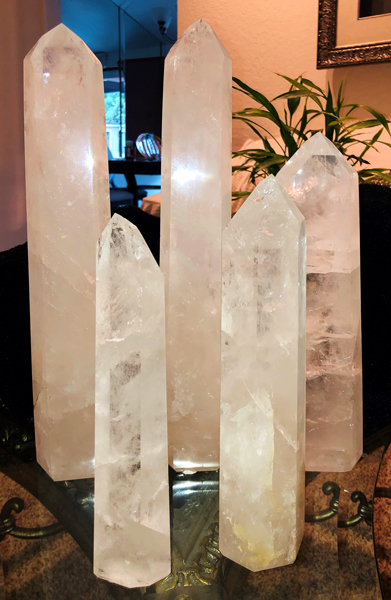 Five tall pillar quartz crystals on display.  They range between 10 and 17 inches in height. Light can be seen shining through them.